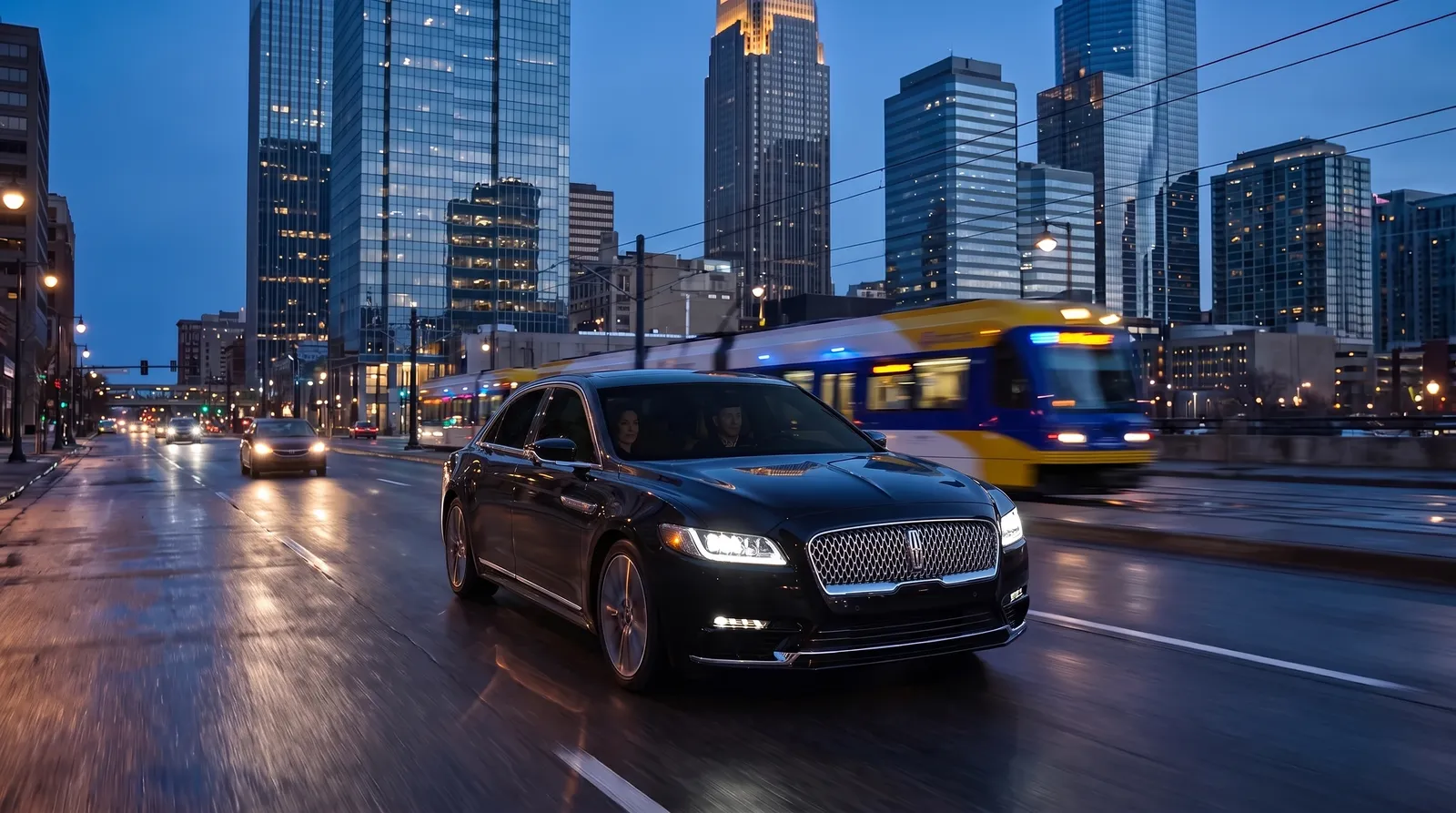 Black Lincoln Continental sedan on a downtown Minneapolis avenue at blue hour with a light-rail train behind